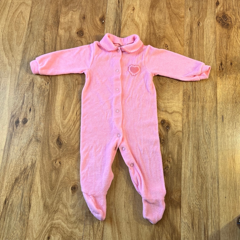May include: A pink baby onesie with a heart-shaped design on the chest. The onesie has a button closure down the front and a collar.
