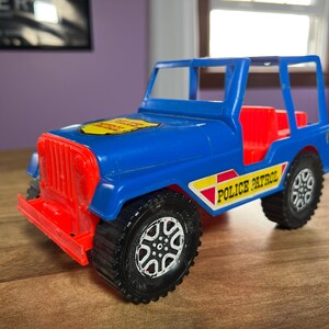 May include: Blue toy jeep with red accents and "Police Patrol" written on the side. The jeep has a yellow sticker on the hood.