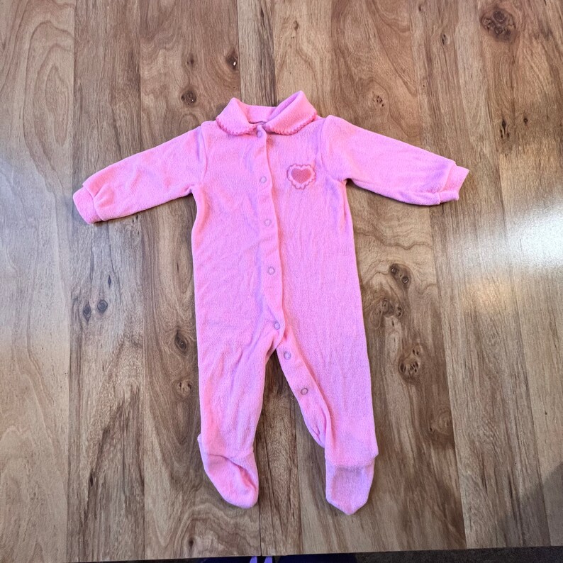 May include: A pink baby onesie with a heart on the chest and button closures.