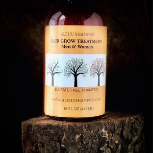 May include: A brown bottle of Alehu Shampoo Hair Grow Treatment, a sulfate-free shampoo. The label is yellow with black text and a graphic of three trees. The bottle is sitting on a wooden surface. 15 fl oz (443 ml).