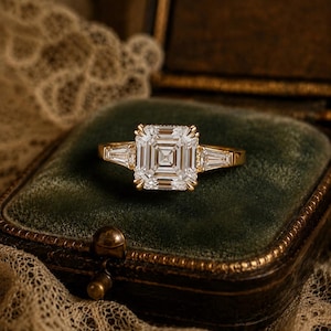 May include: A gold ring featuring a large square-cut center stone with a step-cut facet pattern, flanked by two smaller tapered baguette-cut side stones. The ring is set on a thin gold band, showcasing a classic three-stone design with clear stones.