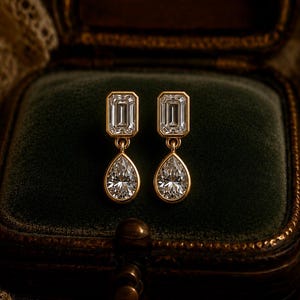 May include: A pair of gold drop earrings featuring a rectangular emerald-cut stone at the top connected to a pear-shaped teardrop stone below. Each earring is set in a polished gold frame, showcasing clear, faceted stones that catch the light.