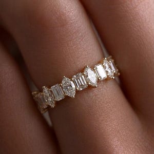May include: A gold band ring featuring an alternating pattern of clear, faceted marquise-cut and emerald-cut stones set in a continuous row. The stones are arranged horizontally around the band, creating a sparkling, geometric design.