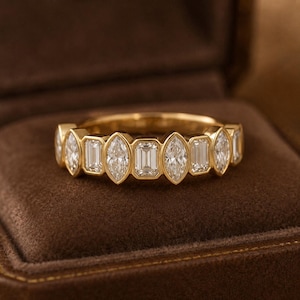 Könnte beinhalten: Ein goldener Ring mit einem abwechselnden Muster aus klaren, im Marquise- und Smaragdschliff facettierten Steinen in Zargenfassung. Der Ring zeigt eine durchgehende Reihe von facettierten Edelsteinen, die den polierten Goldring vollständig umschließen.