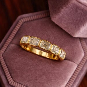 May include: A gold band ring featuring five rectangular-cut, clear gemstones. The ring is set in a gold band and rests on a purple velvet surface, likely a ring box. The gemstones are evenly spaced.