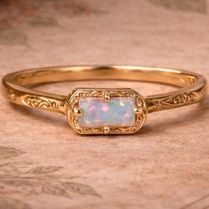 Puede incluir: Un anillo de oro con una piedra de ópalo. El anillo tiene un diseño ornamentado y grabado en la banda y alrededor del ópalo rectangular. El ópalo tiene una mezcla de colores blanco, azul y rosa.