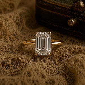 May include: A gold ring featuring a large, rectangular emerald-cut clear gemstone set with four prongs. The band is smooth and polished, showcasing a classic solitaire design with a high-shine finish that reflects light across the faceted stone.