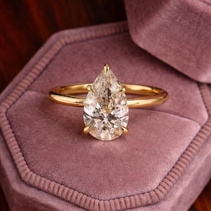 May include: A gold ring featuring a pear-cut diamond. The diamond is clear and faceted, held in place by four prongs. The ring is presented on a pink velvet surface.