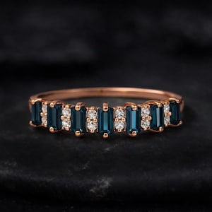 May include: A rose gold ring featuring alternating rectangular blue gemstones and small round white gemstones. The ring is set against a dark background, highlighting the sparkle of the stones.