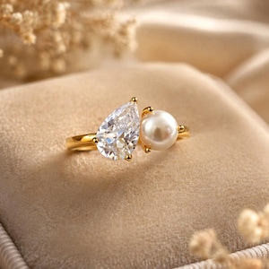 Puede incluir: Un anillo de oro con una gema clara en forma de pera y una perla blanca redonda. El anillo tiene un diseño delicado con una banda de oro y pequeños detalles dorados alrededor de las gemas. El anillo está sobre un fondo suave y neutro.