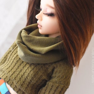 May include: A doll with long brown hair wearing a green knitted jumper with a matching cowl neck scarf.