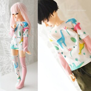 May include: Two dolls wearing matching shirts with a dinosaur print. The shirts are white with pink sleeves and feature a variety of colourful dinosaurs, leaves, and flowers. The dolls are wearing different coloured socks.