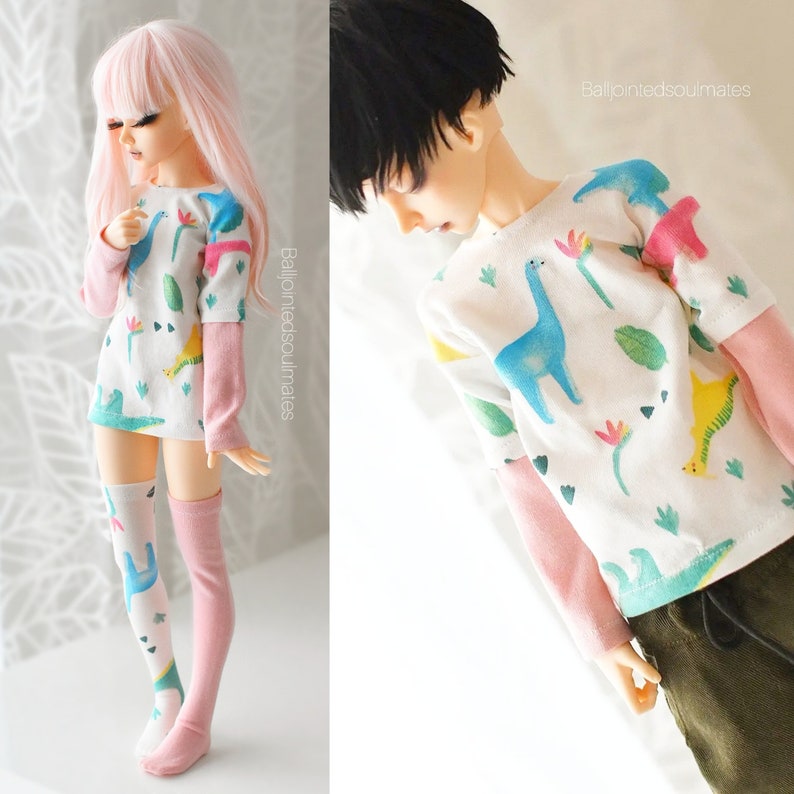 May include: Two dolls wearing matching shirts with a dinosaur print. The shirts are white with pink sleeves and feature a variety of colourful dinosaurs, leaves, and flowers. The dolls are wearing different coloured socks.
