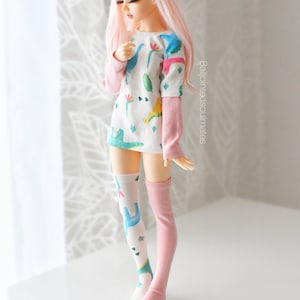 May include: A pink haired doll wearing a white long-sleeved shirt with a dinosaur print and pink and white knee-high socks.