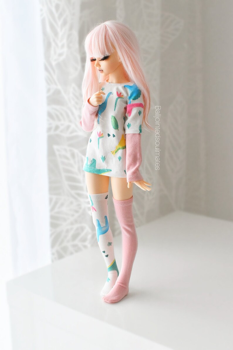 May include: A pink haired doll wearing a white long-sleeved shirt with a dinosaur print and pink and white knee-high socks.