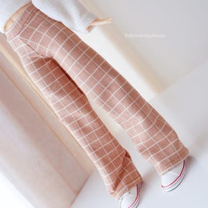 May include: A doll wearing a white sweater and tan plaid pants with white sneakers. The pants have a grid pattern in white. The sneakers have red and pink accents. The doll's outfit is set against a white background.