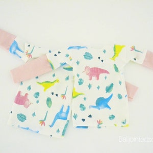 May include: Two white long-sleeved shirts with pink trim on the sleeves. The shirts have a white background with a pattern of colourful dinosaurs and green leaves.
