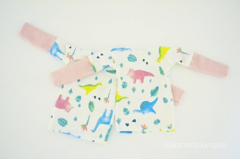 May include: Two white long-sleeved shirts with pink trim on the sleeves. The shirts have a white background with a pattern of colourful dinosaurs and green leaves.