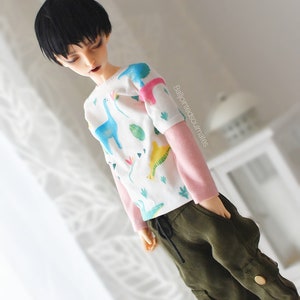 May include: A doll wearing a white long-sleeved shirt with a dinosaur print and pink sleeves. The doll is wearing green cargo trousers with a drawstring waist.
