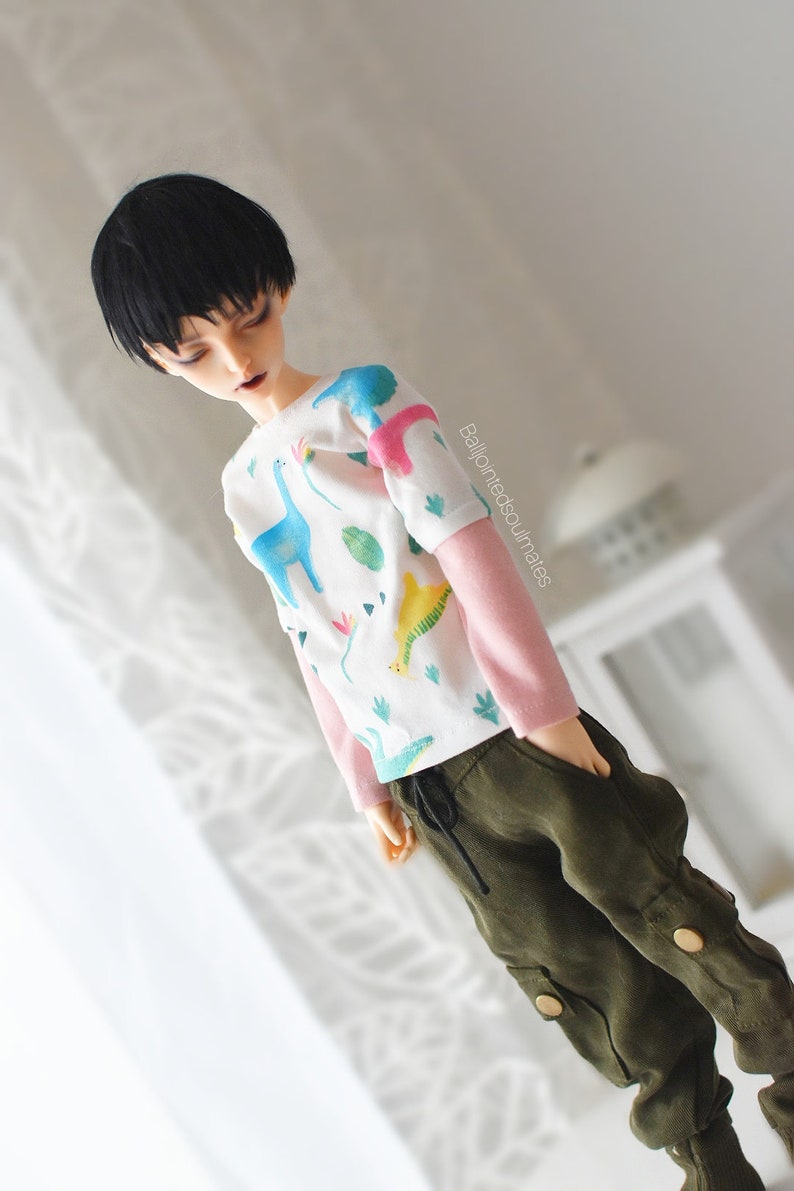 May include: A doll wearing a white long-sleeved shirt with a dinosaur print and pink sleeves. The doll is wearing green cargo trousers with a drawstring waist.
