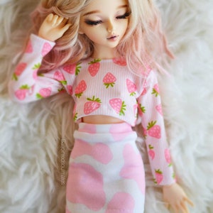 Peut inclure: Une poup&eacute;e portant un haut court &agrave; imprim&eacute; fraises rose et blanc et une jupe &agrave; pois rose et blanc. La poup&eacute;e a de longs cheveux blonds avec des reflets roses et est allong&eacute;e sur une surface blanche et moelleuse.