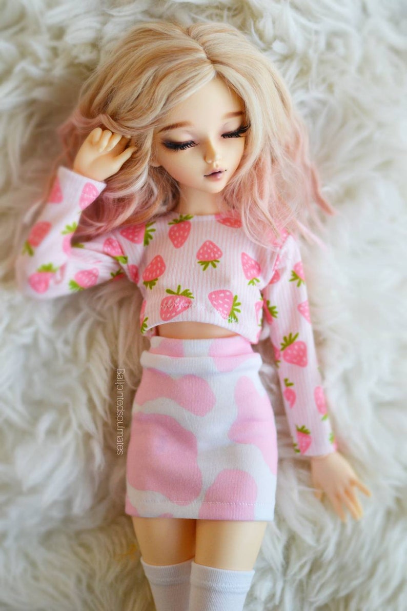 Peut inclure: Une poup&eacute;e portant un haut court &agrave; imprim&eacute; fraises rose et blanc et une jupe &agrave; pois rose et blanc. La poup&eacute;e a de longs cheveux blonds avec des reflets roses et est allong&eacute;e sur une surface blanche et moelleuse.