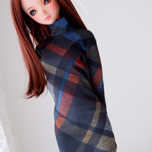 Könnte beinhalten: Eine Puppe mit langen, glatten, rotbraunen Haaren, die ein schwarzes, blaues und braunes kariertes Rollkragenkleid trägt. Die Puppe trägt weiße Socken und hat einen neutralen Gesichtsausdruck.