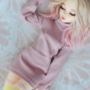 Puede incluir: Una muñeca con el pelo largo y rubio con reflejos rosas que lleva un jersey rosa de cuello alto y leotardos amarillos. La muñeca está tumbada sobre una superficie blanca y esponjosa.