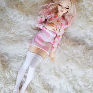 Peut inclure: Une poup&eacute;e rose et blanche aux longs cheveux blonds portant un haut et une jupe &agrave; imprim&eacute; fraises rose et blanc avec des chaussettes blanches montantes. La poup&eacute;e est allong&eacute;e sur une surface blanche et moelleuse.