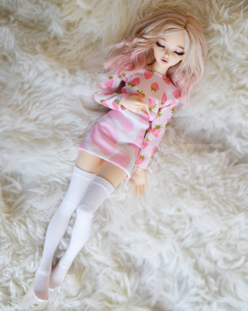 Peut inclure: Une poup&eacute;e rose et blanche aux longs cheveux blonds portant un haut et une jupe &agrave; imprim&eacute; fraises rose et blanc avec des chaussettes blanches montantes. La poup&eacute;e est allong&eacute;e sur une surface blanche et moelleuse.