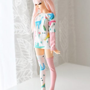 May include: A pink-haired doll wearing a white long-sleeved shirt with a dinosaur print and pink knee-high socks. The shirt has a green, blue, and yellow dinosaur print. The doll is standing on a white surface.