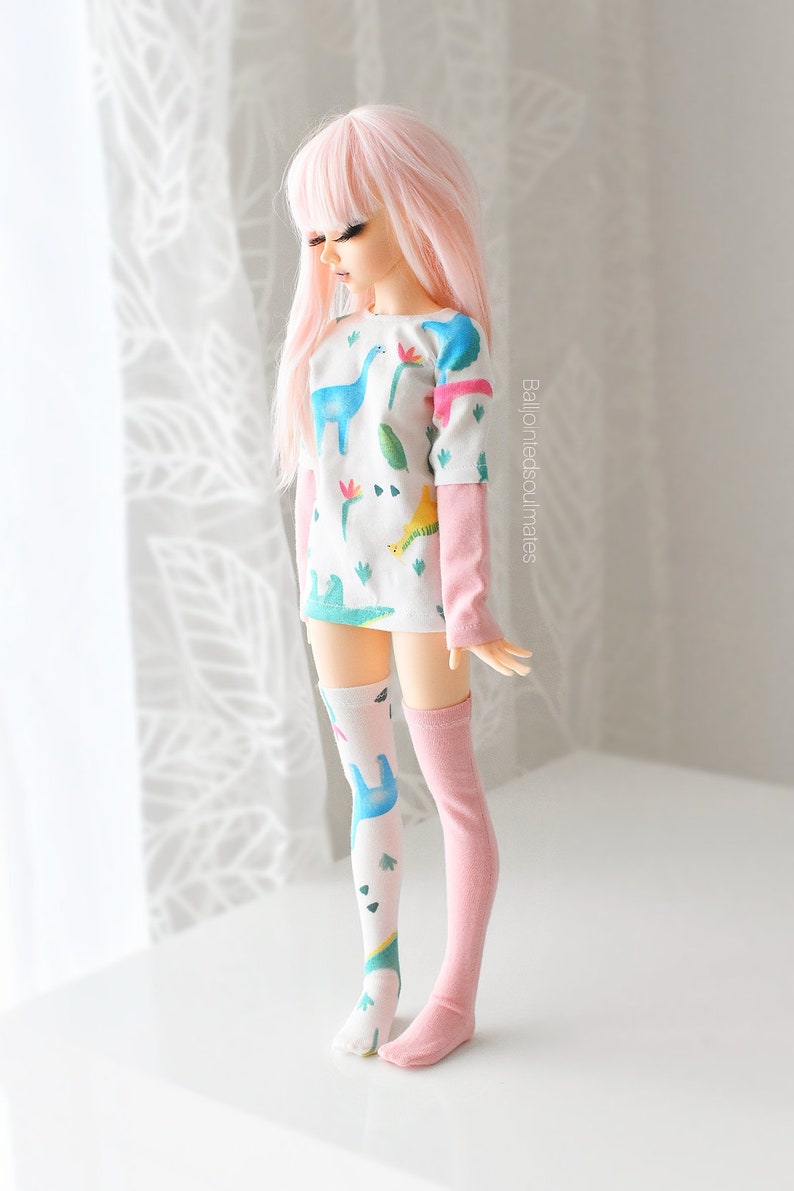 May include: A pink-haired doll wearing a white long-sleeved shirt with a dinosaur print and pink knee-high socks. The shirt has a green, blue, and yellow dinosaur print. The doll is standing on a white surface.