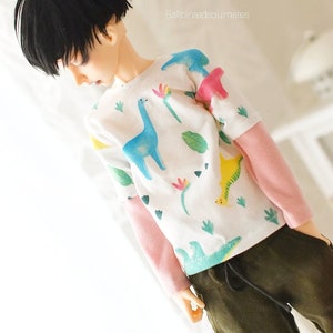 May include: A doll wearing a white long-sleeved shirt with a colourful dinosaur print and pink sleeves. The doll is also wearing dark green trousers with a drawstring.
