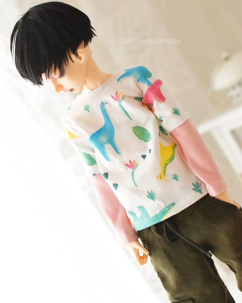 May include: A doll wearing a white long-sleeved shirt with a colourful dinosaur print and pink sleeves. The doll is also wearing dark green trousers with a drawstring.
