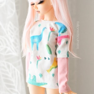 May include: A pink haired doll wearing a white t-shirt with a dinosaur print and pink long sleeves. The doll is wearing white and pink knee-high socks.