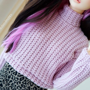 Può includere: Un maglione corto rosa chiaro a collo alto con maniche lunghe. Il maglione è realizzato in tessuto a maglia grossa. La bambola indossa una gonna con stampa leopardata nera e marrone. La bambola ha i capelli scuri con riflessi rosa. Il testo "balljointedsoulmates" è visibile.