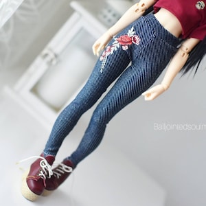 Pode incluir: Uma boneca vestindo um top cropped bordô e leggings com aparência de jeans com bordado floral. A boneca também está usando tênis bordô com cadarços brancos. O fundo é branco.