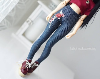 Peut inclure: Une poupée vêtue d'un crop top bordeaux et d'un legging en jean avec une broderie florale. La poupée porte également des baskets bordeaux avec des lacets blancs. Le fond est blanc.