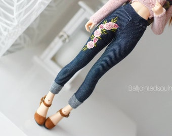 Peut inclure: Un pantalon en jean pour poupée avec broderie florale. Le jean est de couleur bleu foncé avec un revers et présente un motif floral rose et vert sur la jambe gauche. La poupée porte également un pull rose et des chaussures marron.