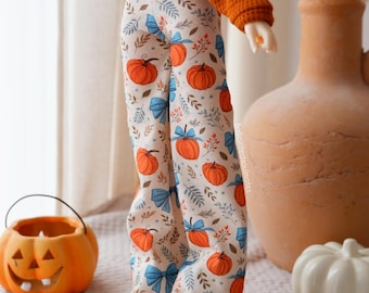 Könnte beinhalten: Eine Puppe trägt ein langärmeliges Oberteil aus orangefarbenem Waffelstrick und eine cremefarbene weite Hose mit einem Muster aus orangefarbenen Kürbissen, blauen Schleifen und grünem Blattwerk. Die Puppe trägt beigefarbene Slipper.