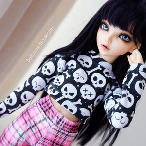 Könnte beinhalten: Eine Puppe mit langen schwarzen Haaren, die ein schwarzes und weißes Oberteil mit Totenkopf-Muster und einen rosa-schwarzen karierten Rock trägt.