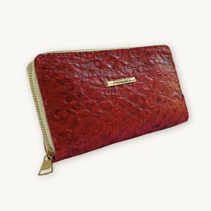May include: A rectangular red wallet with a textured surface and a gold-toned zipper closure. A small, horizontal gold-colored metal plate is attached to the front, featuring the engraved text Ambani in a clear, sans-serif font.
