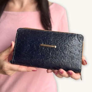 May include: A rectangular navy blue wallet with a textured, crinkled surface finish. The wallet features a gold-toned metal zipper along the top edge and a small, horizontal gold-toned metal plaque centered on the front with the text Ambar.