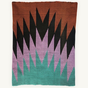 Puede incluir: Un tapiz textil rectangular tejido con un patrón geométrico de puntas en zigzag verticales y afiladas. El diseño transita desde una base de color verde azulado profundo en la parte inferior hasta lavanda, negro y marrón terracota.