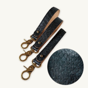 Pode incluir: Três alças de pulso pretas texturizadas com o verso em tom natural, cada uma com um mosquetão giratório de metal cor de latão e um rebite de latão a condizer. Uma inserção circular mostra um close-up da textura escura e escamosa.