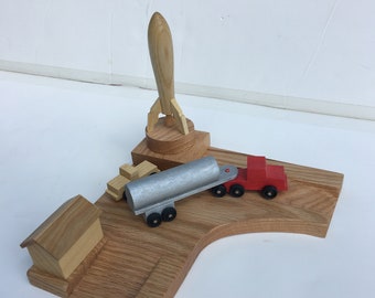 May include: A wooden toy set featuring a rocket, a truck, and a house. The rocket is made of light wood and has a pointed nose. The truck is red and has a silver tank. The house is made of light wood and has a simple design. The toys are arranged on a wooden base.