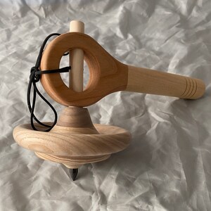 May include: A wooden spinning top with a string and a wooden handle. The top is made of light wood and has a dark wood center. The handle is also made of light wood and has a dark wood ring around the center.