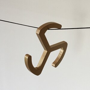 May include: A gold-colored metal hook with a unique, abstract design. The hook is hanging from a black string.