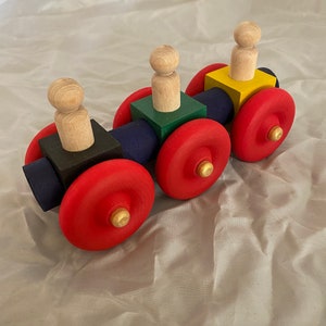 May include: A wooden toy train with three carriages, each with a different coloured block and a wooden person on top. The train has red wheels and is on a white fabric surface.