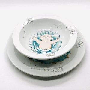 May include: A white ceramic baby bowl and plate set with a blue cabbage and a baby inside. The bowl and plate are decorated with small white bunnies.